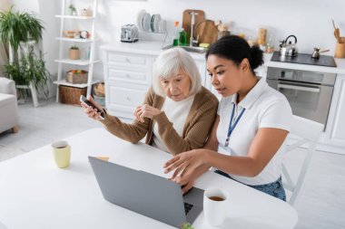 multiracial social worker using laptop near retired woman with smartphone  clipart