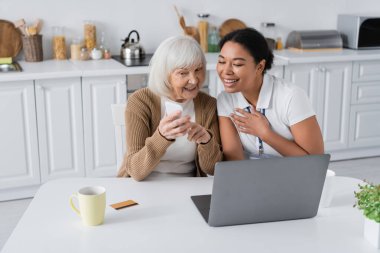 Neşeli çokkültürlü sosyal hizmetler görevlisi dizüstü bilgisayarın yanındaki emekli kadının elinde akıllı telefona bakıyor. 