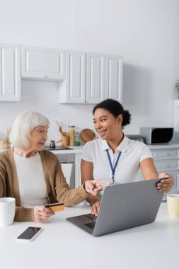Mutlu çok ırklı sosyal hizmetler görevlisinin yanındaki dizüstü bilgisayarı gösteren neşeli yaşlı kadın.