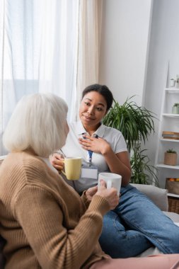 sentimental multiracial social worker chatting with senior woman while having tea in living room  clipart