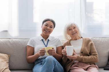 cheerful multiracial social worker having tea with senior woman in living room  clipart