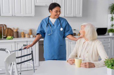 Steteskoplu neşeli hemşire elinde çayıyla mutfakta oturan mutlu yaşlı kadına bakıyor. 
