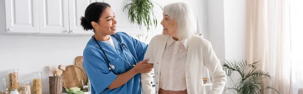 Ευτυχισμένη Πολυφυλετική Νοσοκόμα Μπλε Στολή Κοιτάζοντας Ηλικιωμένη Γυναίκα Γκρίζα Μαλλιά — Φωτογραφία Αρχείου