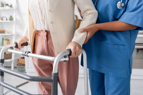 обрезанный вид многорасовой медсестры в синей форме, поддерживающей пожилую женщину, идущую с помощью ходячего дома