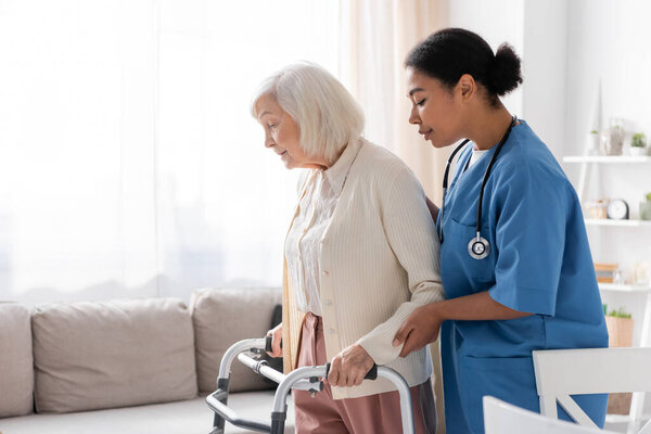 brunette multiracial nurse in uniform supporting retired woman with grey hair walking with walker at home 