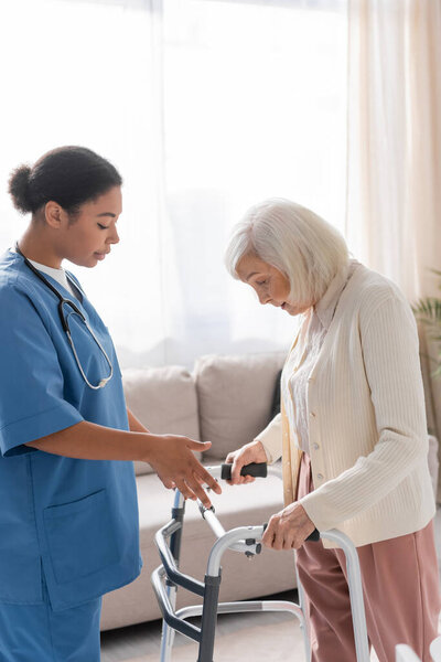 брюнетка многорасовая медсестра в синей форме смотрит на пожилую женщину с седыми волосами ходьба с помощью ходунков на дому 
