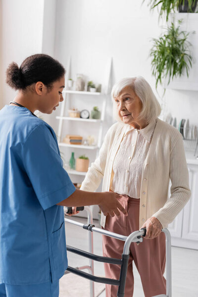 senior woman with grey hair walking with help of walker and looking at brunette multiracial nurse 
