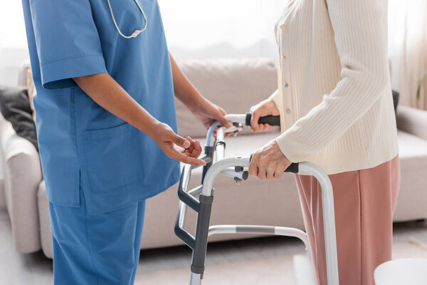 обрезанный вид мультирасового сиделки в синей форме, поддерживающей пожилую женщину, идущую с помощью ходячего дома