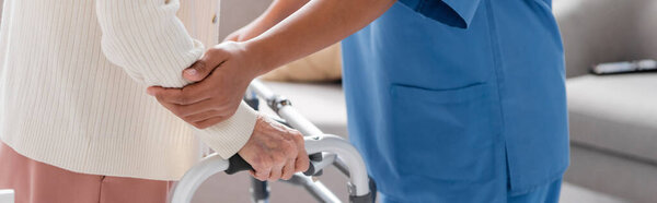 cropped view of multiracial caregiver in blue uniform comforting senior woman walking with help of walker at home, banner 