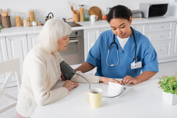 表にお茶のカップの横に白髪の老女の血圧を測定する多人種看護師 — ストック写真
