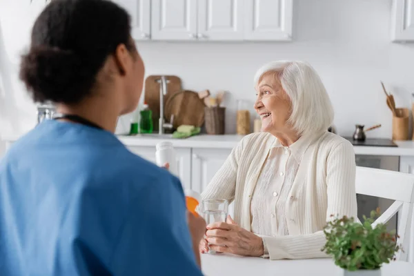 白髪の幸せな引退した女性は前景がぼやけて多人種間の看護師の近くを見て — ストック写真