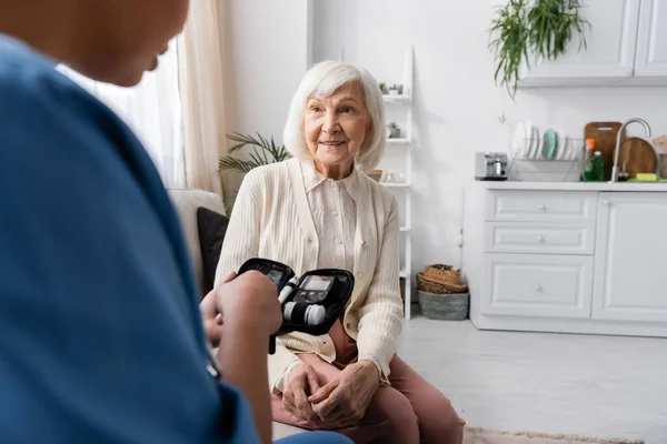 快乐的老年妇女坐在沙发上 看着持有糖尿病药包的多种族护士 — 图库照片