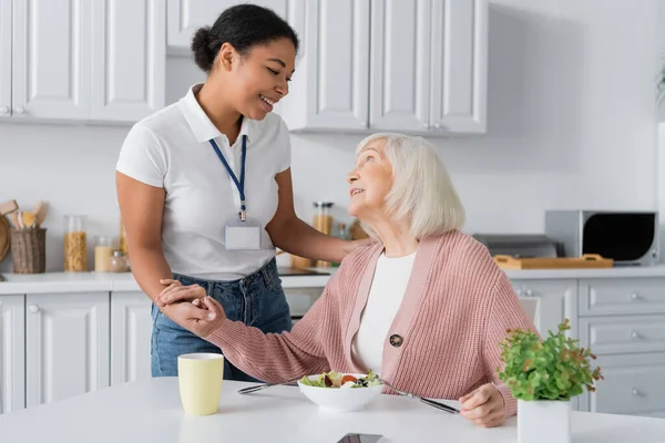 Fröhliche Multirassische Sozialarbeiterin Hält Die Hände Einer Dankbaren Rentnerin — Stockfoto