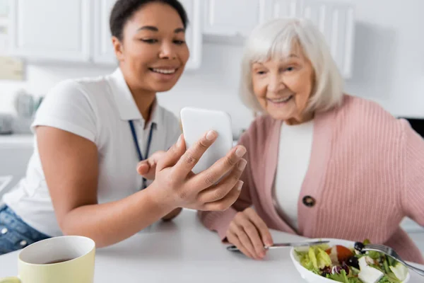 Fröhlicher Multiethnischer Sozialarbeiter Zeigt Seniorin Beim Mittagessen Küche Smartphone — Stockfoto