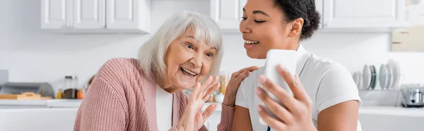 スマートフォンを持ちながらビデオ通話中に手を振っている高齢女性の陽気な多人種社会労働者 バナー — ストック写真