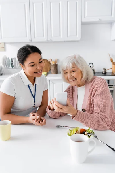 Šťastný Senior Žena Pomocí Smartphone Blízkosti Multiracial Sociální Pracovník Doma — Stock fotografie