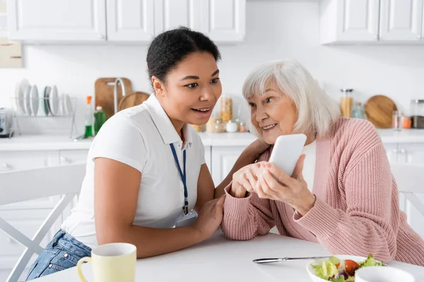 快乐的老年妇女在家里给多种族的社会工作者展示智能手机 让他们大吃一惊 — 图库照片