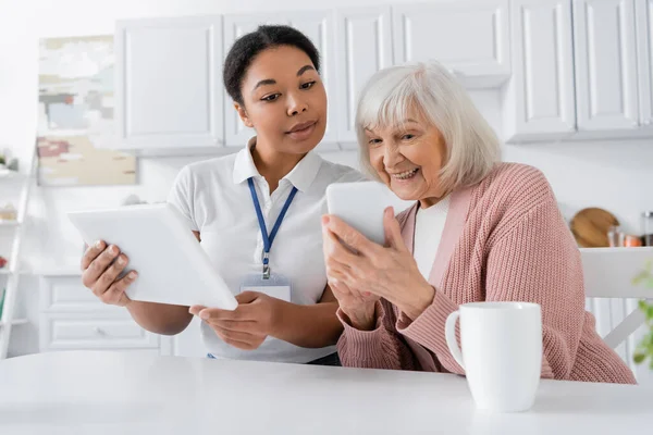 brunette multiracial social worker holding digital tablet near senior woman with smartphone in kitchen