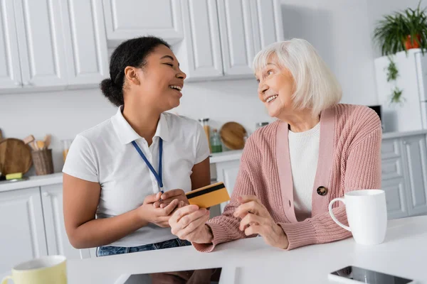 happy multiracial social worker holding credit card near senior woman at home