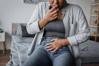 Cropped view of multiracial woman touching belly while sitting on bed in bedroom  clipart