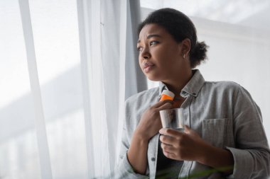 Depresyonda olan üzgün, çok ırklı bir kadın. Elinde haplar ve pencerenin yanında su tutuyor. 