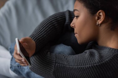 Yatak odasında boş ekranlı akıllı telefon tutan depresif çok ırklı kadının yan görüntüsü 