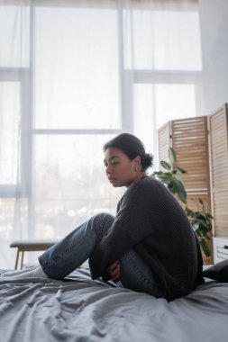 Evde yatakta örgü kazaklı yalnız bir kadın oturuyor. 