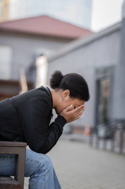 Kentsel caddede bankta otururken yüzünü örten depresyondaki çok ırklı kadın. 