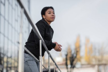 multiracial woman with mental problem looking away while standing near railing on street  clipart