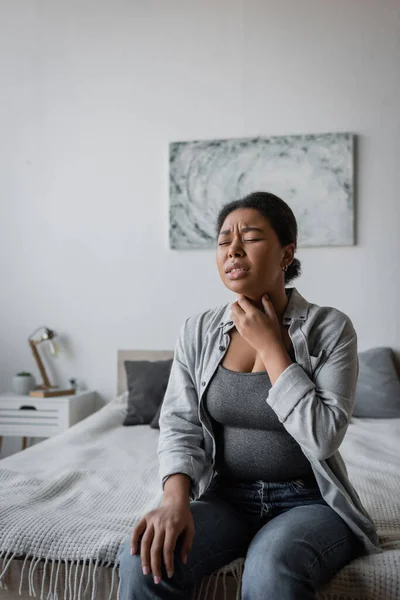 Στρεσαρισμένη Πολυφυλετική Γυναίκα Που Αγγίζει Πονόλαιμο Ενώ Κάθεται Στο Κρεβάτι — Φωτογραφία Αρχείου