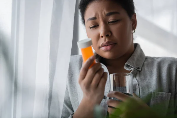 Sad multiracial woman holding blurred pills and water near curtain at home