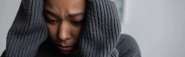 Worried multiracial woman in sweater touching head at home, banner