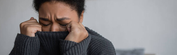Депрессивная многорасовая женщина в трикотажном свитере плачет дома, баннер 