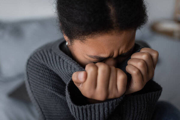 Sad multiracial woman in sweater crying in blurred bedroom at home 