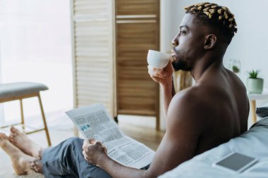 Side view of african american man with vitiligo holding coffee and newspaper in bedroom  clipart