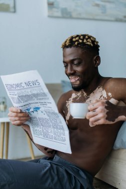 Gülümseyen Afro-Amerikan adam Vitiligo ile ekonomi gazetesi okuyor ve yatak odasında kahve tutuyor. 