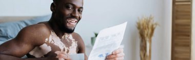 Gülümseyen Afrikalı Amerikalı Vitiligo 'lu adam yatak odasında kahve ve gazete tutuyor. 