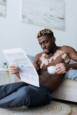 Muscular african american man with vitiligo holding coffee and reading newspaper in bedroom  clipart