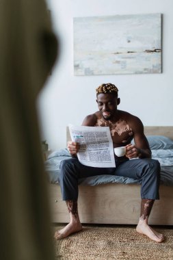 Smiling african american man with vitiligo reading news and holding coffee on bed at home  clipart