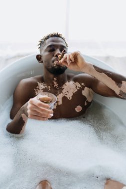 Vitiligo 'lu Afrikalı Amerikalı bir adam puro içiyor ve evde banyo yaparken viski tutuyor. 
