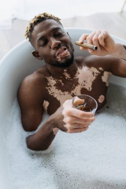Vitiligo 'lu, elinde puro ve viskiyle evde köpükle banyo yapan Afro-Amerikan bir adamın yüksek açılı görüntüsü. 