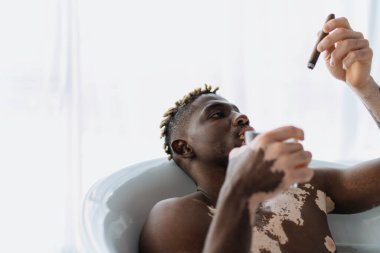 Vitiligo 'lu genç Afro-Amerikalı adam evde banyo yaparken puroya bakıyor ve cam tutuyor. 
