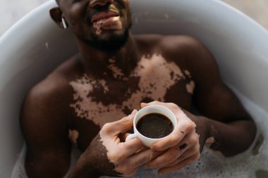 Vitiligo 'su olan gülümseyen Afro-Amerikan bir adamın küvet kulaklığı ve kahve fincanı kullanırken banyo yaparken görüntüsü. 
