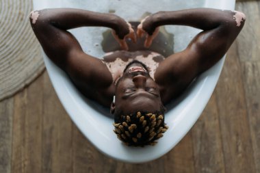Overhead view of african american man with vitiligo listening music and taking bath at home  clipart