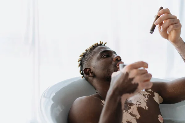 stock image Young african american man with vitiligo looking at cigar and holding glass while taking bath at home 