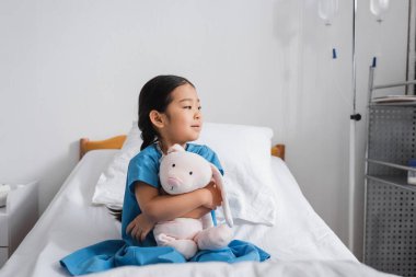 Düşünceli Asyalı kız oyuncak tavşanı kucaklıyor ve hastane yatağında otururken gözlerini kaçırıyor.