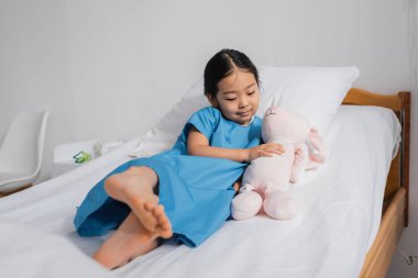 Pediyatri kliniğinde yatarken oyuncak tavşanı kucaklayan hastane önlüklü çıplak ayaklı Asyalı kız.
