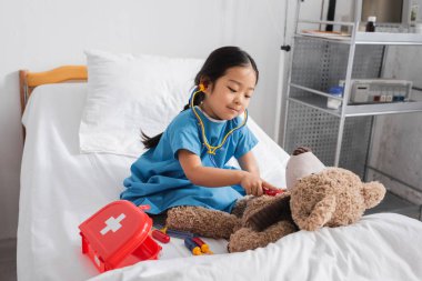 Oyuncak steteskoplu pozitif Asyalı kız pediatri kliniğinde ayıcığı yatakta muayene ediyor.
