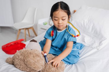 Hastane önlüklü Asyalı çocuk klinikte oynarken oyuncak şırıngayla oyuncak ayıya iğne yapıyor.