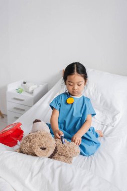 Hastane önlüklü Asyalı çocuk, klinikte yatakta oynarken oyuncak şırıngayla oyuncak ayıya iğne yapıyor.